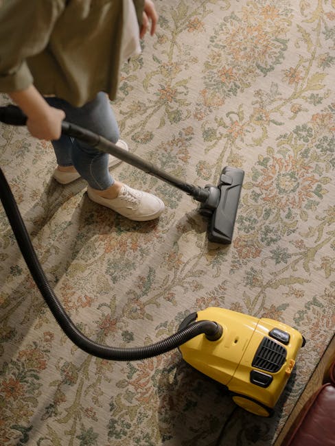A professional cleaning technician wearing full protective gear including a white suit, gloves, and a mask, performs deep carpet cleaning on a light beige carpet in a well-lit residential room. The te
