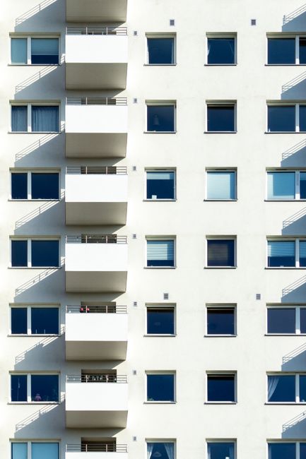 A close-up view of the exterior of a modern, multi-story residential building with white stucco walls and large rectangular windows featuring white frames. The windows are fitted with sheer curtains, 