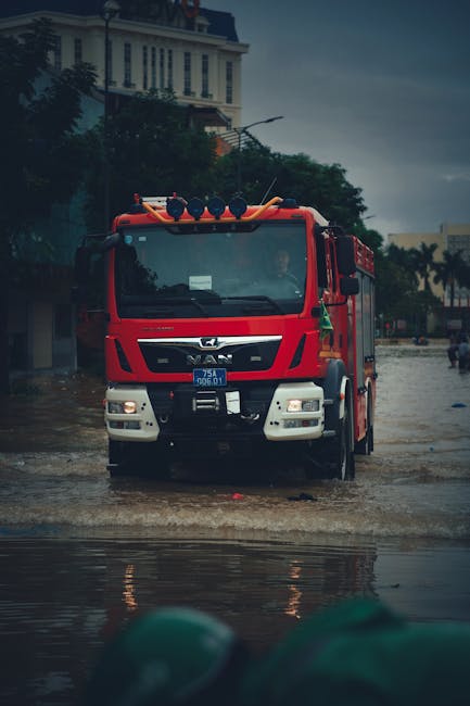 A red fire truck from St Helier Carpet Cleaners is positioned on a flooded street during heavy rainfall, with water pooling around the vehicle's wheels. The truck has multiple blue flashing lights on 