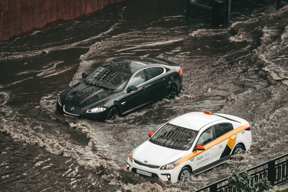 A red fire truck from St Helier Carpet Cleaners is positioned on a flooded street during heavy rainfall, with water pooling around the vehicle's wheels. The truck has multiple blue flashing lights on 